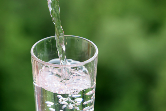 Pouring clean water into drinking glass on green nature background. Concept of health and freshness, thirst, water purification